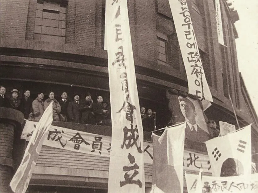 큰 현수막과 깃발이 걸린 건물 앞 연단에 사람들이 모인 흑백 사진