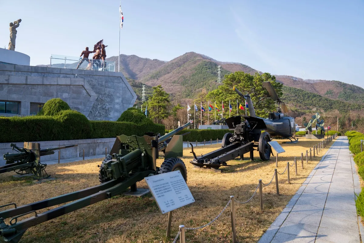 야외 전시장에 놓인 대포와 전투 장비, 뒤로 산이 보이는 풍경